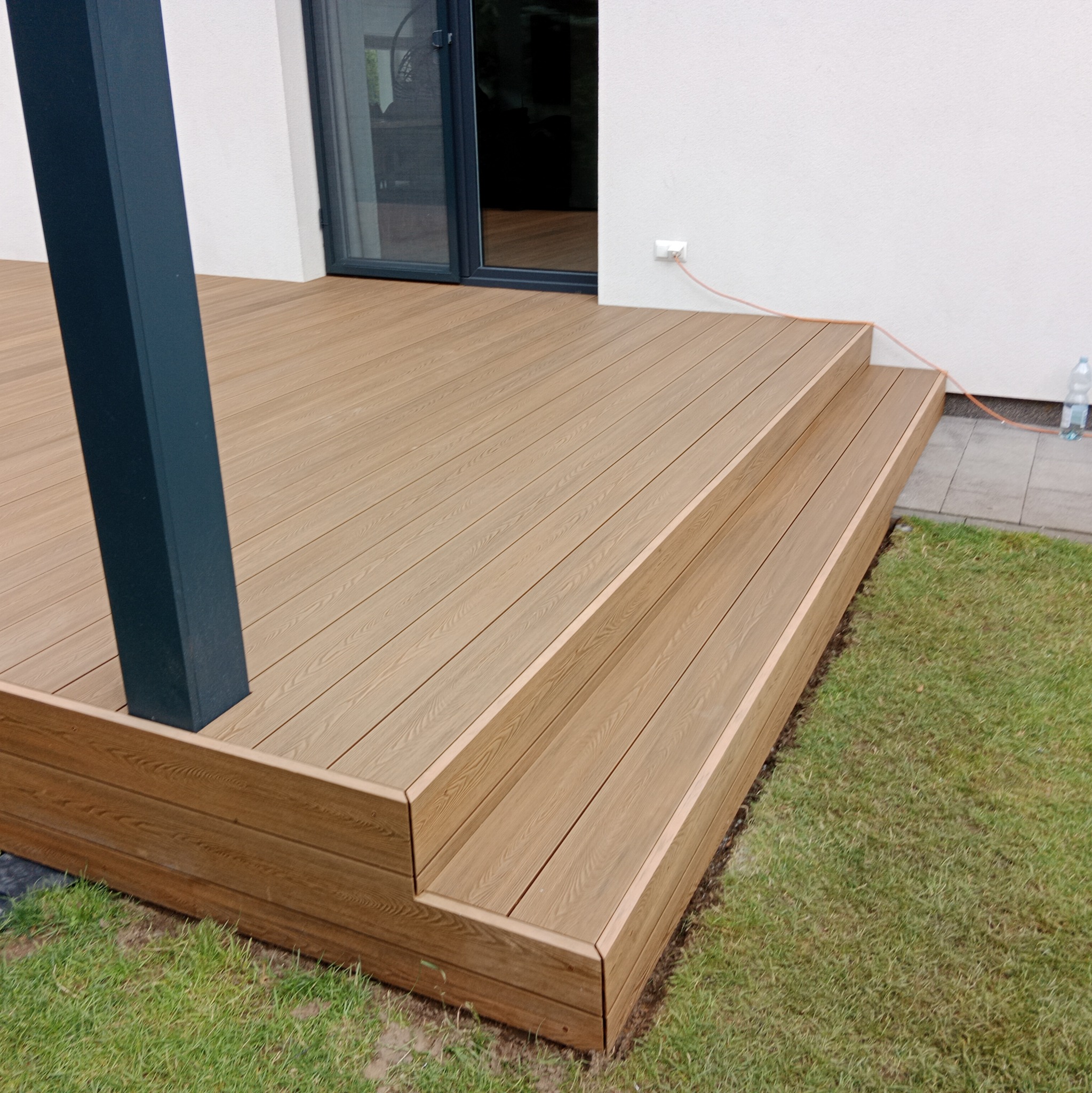 Nowoczesny taras z desek kompozytowych w odcieniach brązu, z dwoma stopniami, czarną podporą i widokiem na trawnik oraz ścianę domu z oknem przesuwnym i gniazdkiem elektrycznym.