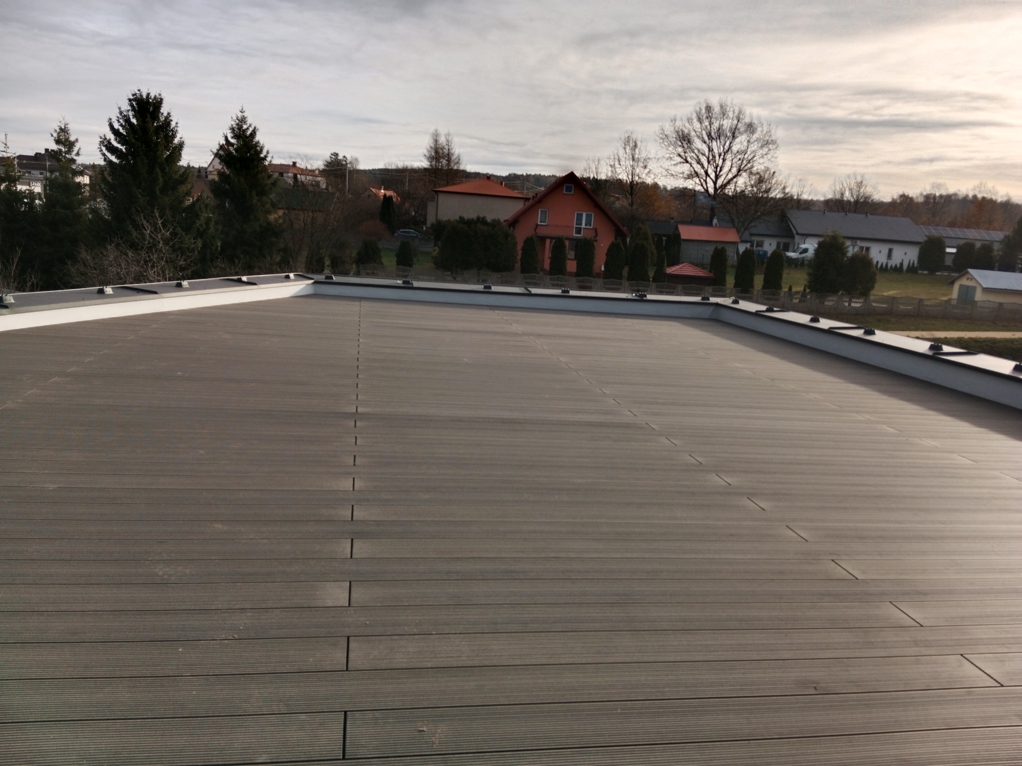 Rozległy, brązowy taras z desek kompozytowych na dachu budynku, z widokiem na zabudowę jednorodzinną i drzewa w tle, wykonany w pochmurny dzień.