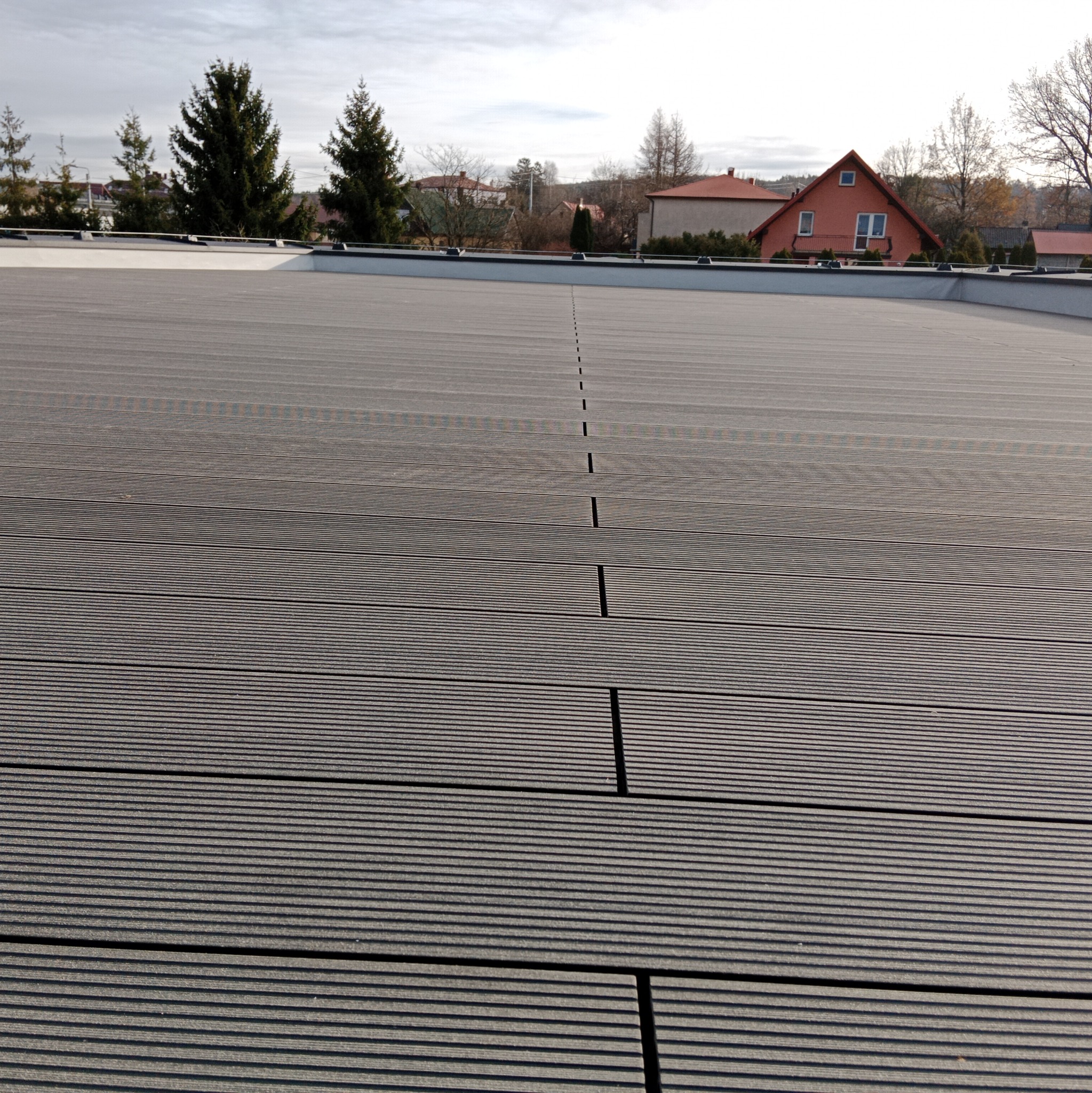 Szary taras z desek kompozytowych, widok z góry na ułożoną powierzchnię, z widocznymi szczelinami dylatacyjnymi i budynkami w tle.