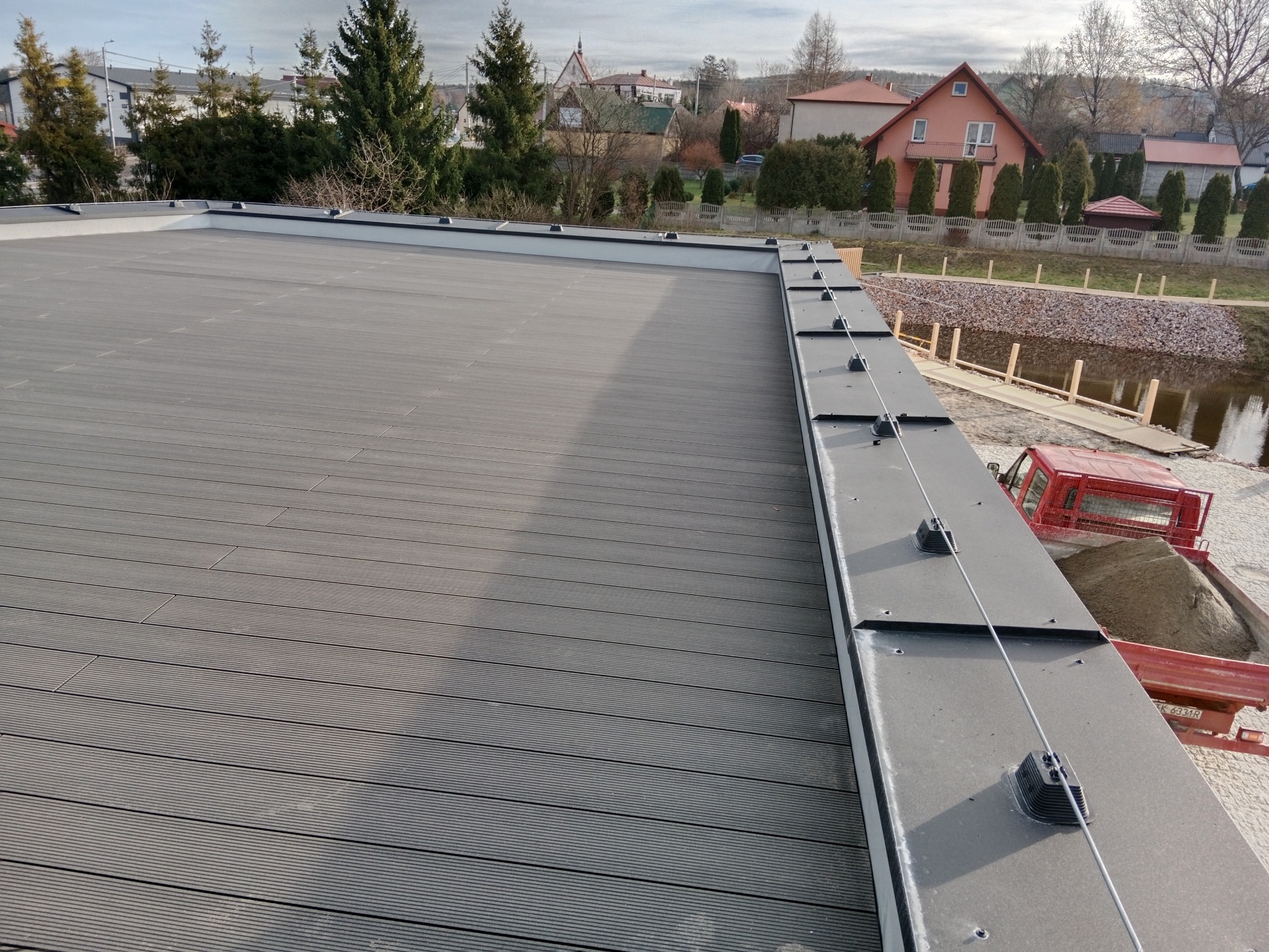 Szary taras z desek kompozytowych z widocznym systemem odgromowym, w tle zabudowa jednorodzinna i rzeka.