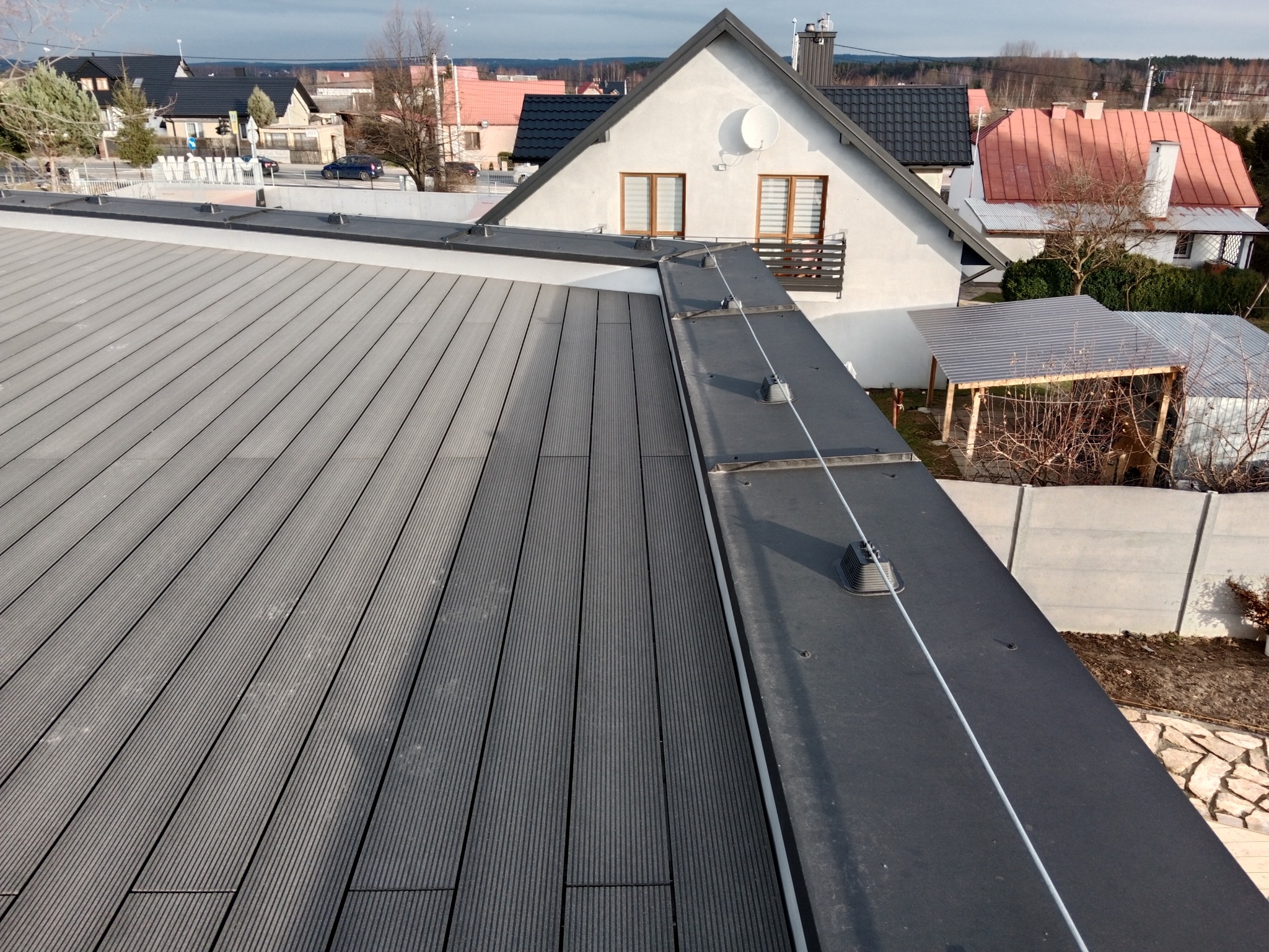 Szary taras z desek kompozytowych na dachu budynku, widoczne elementy mocujące i linka zabezpieczająca, w tle zabudowa jednorodzinna.