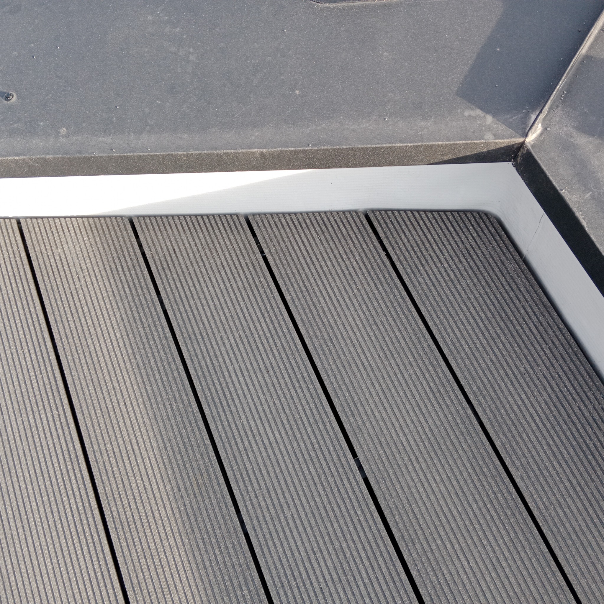 Fragment tarasu kompozytowego w odcieniach szarości, z widoczną białą listwą wykończeniową przy ścianie i czarnymi fugami.