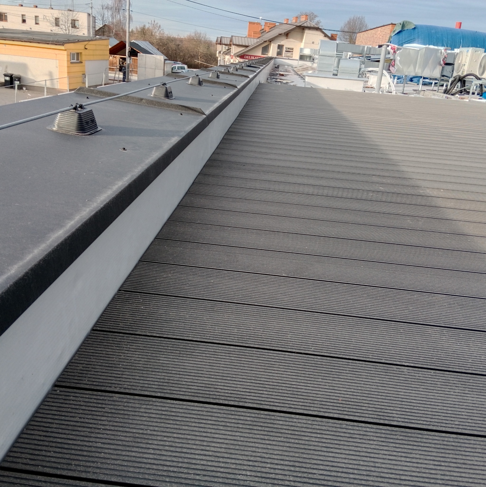 Szare deski tarasowe z kompozytu ułożone na dachu budynku, widoczne elementy instalacji odgromowej i budynki w tle.