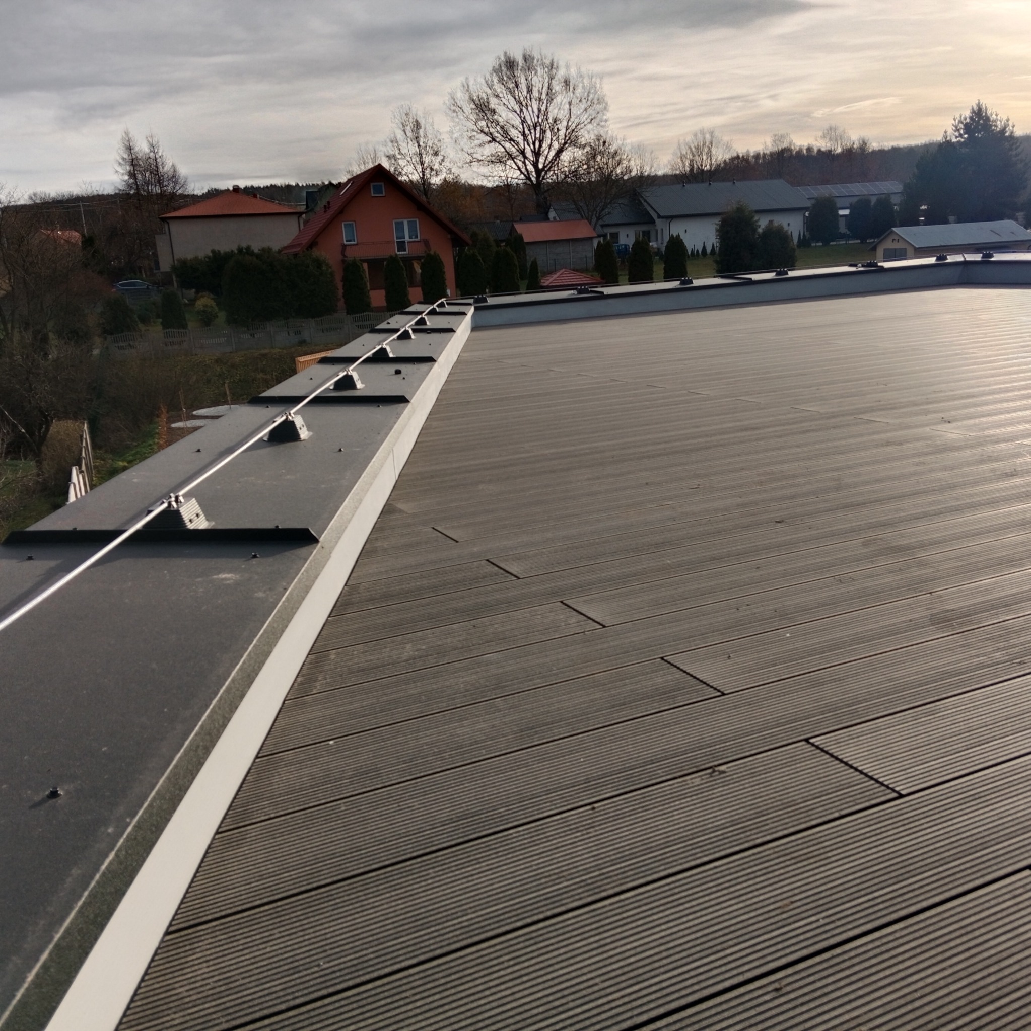 Nowoczesny taras z desek kompozytowych w kolorze grafitowym, zabezpieczony balustradą z odgromnikiem, widok na domy jednorodzinne w tle.