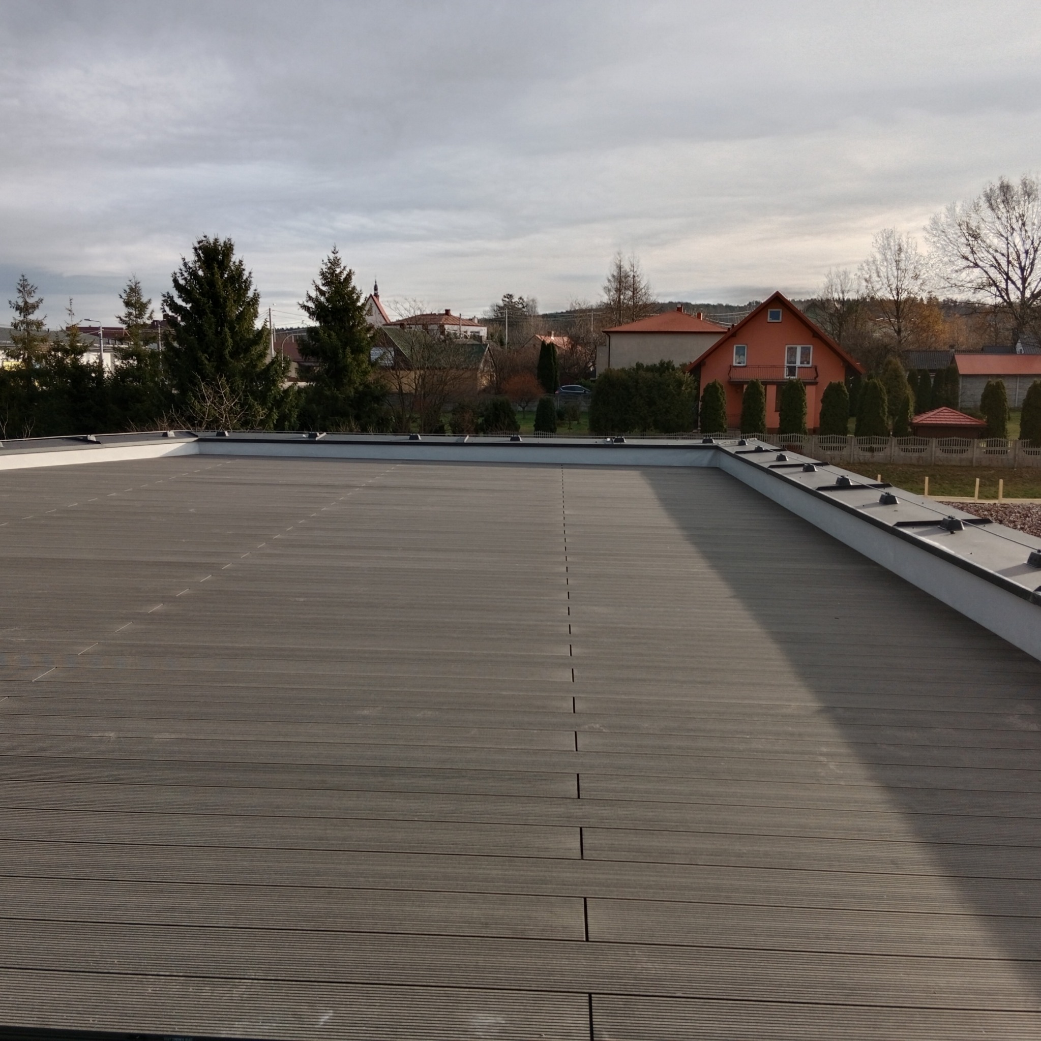 Szary taras kompozytowy na dachu budynku z widocznymi łączeniami desek i elementami wykończeniowymi, w tle zabudowa jednorodzinna.