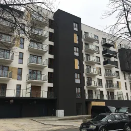 Nowoczesny budynek mieszkalny z balkonami zabezpieczonymi szklanymi balustradami, widok z poziomu ulicy, samochody zaparkowane przed budynkiem.