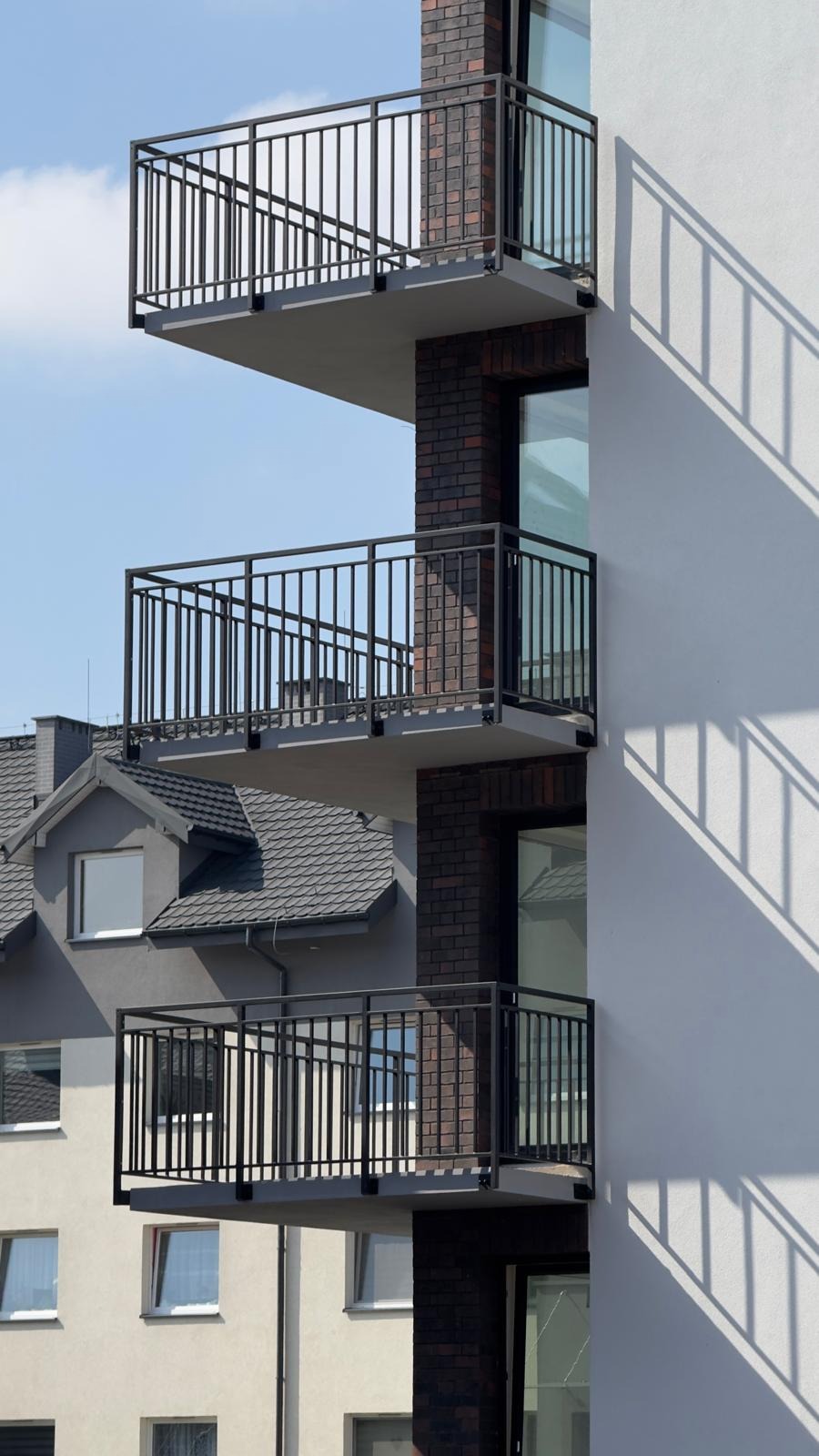 Trzy balkony z czarnymi, metalowymi balustradami na nowoczesnym budynku z ceglanymi detalami i białymi ścianami, cień balustrady na fasadzie.