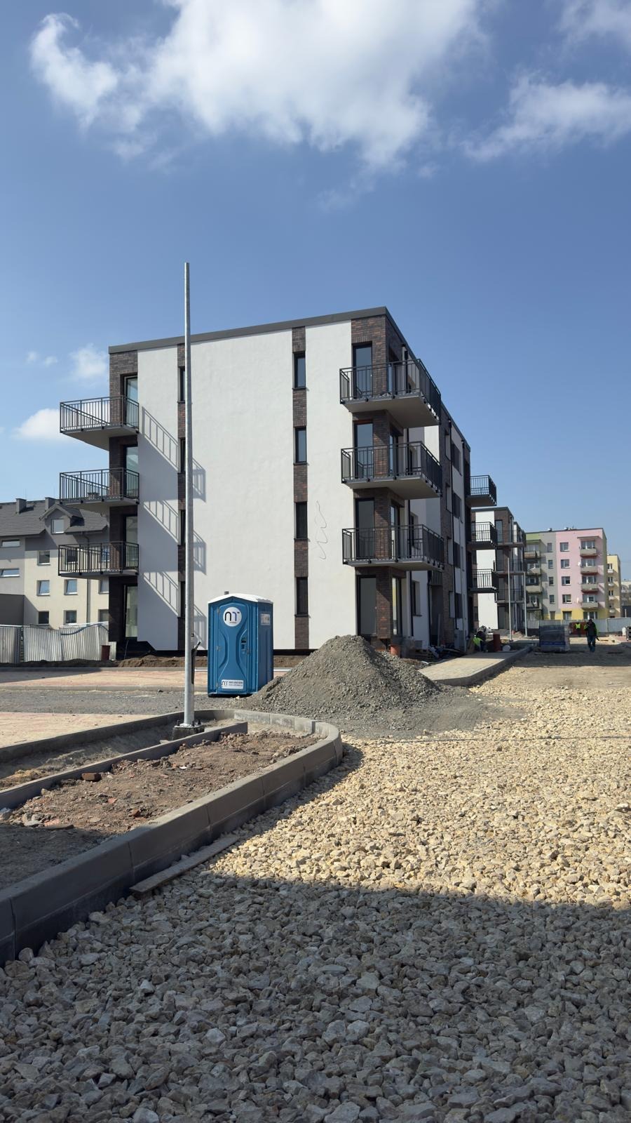 Nowe budynki mieszkalne z balkonami z czarnymi balustradami, widok z placu budowy z przenośną toaletą i stertą gruzu.