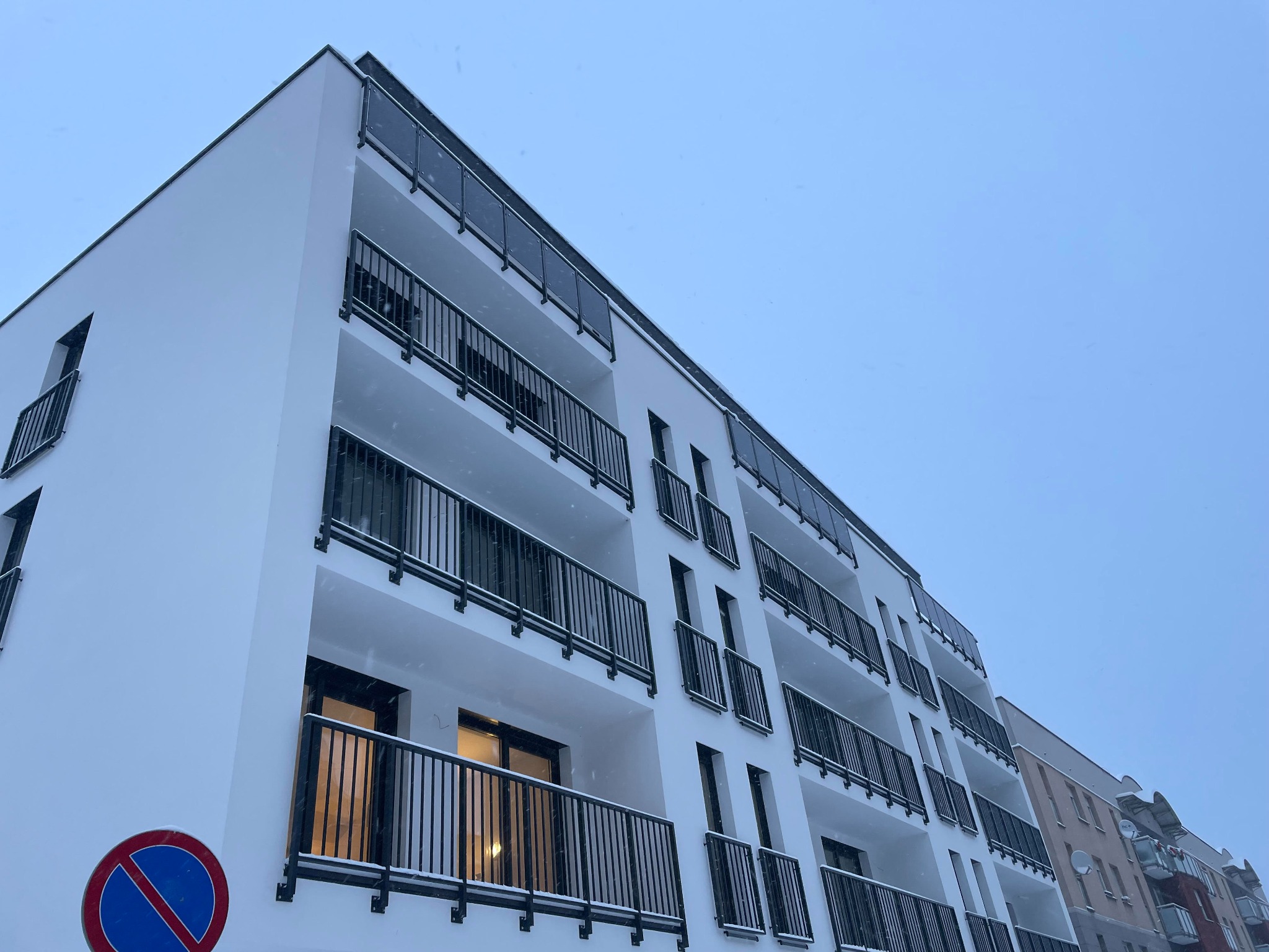 Nowoczesny budynek mieszkalny z białymi elewacjami i balkonami z czarnymi balustradami, widziany pod kątem w pochmurny dzień, z widocznym znakiem zakazu parkowania.