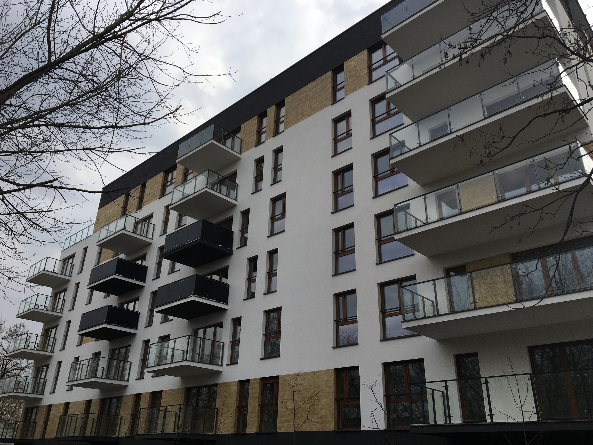 Nowoczesny budynek mieszkalny z balkonami z balustradami szklanymi i metalowymi, widok z dołu na tle pochmurnego nieba i drzew bez liści.