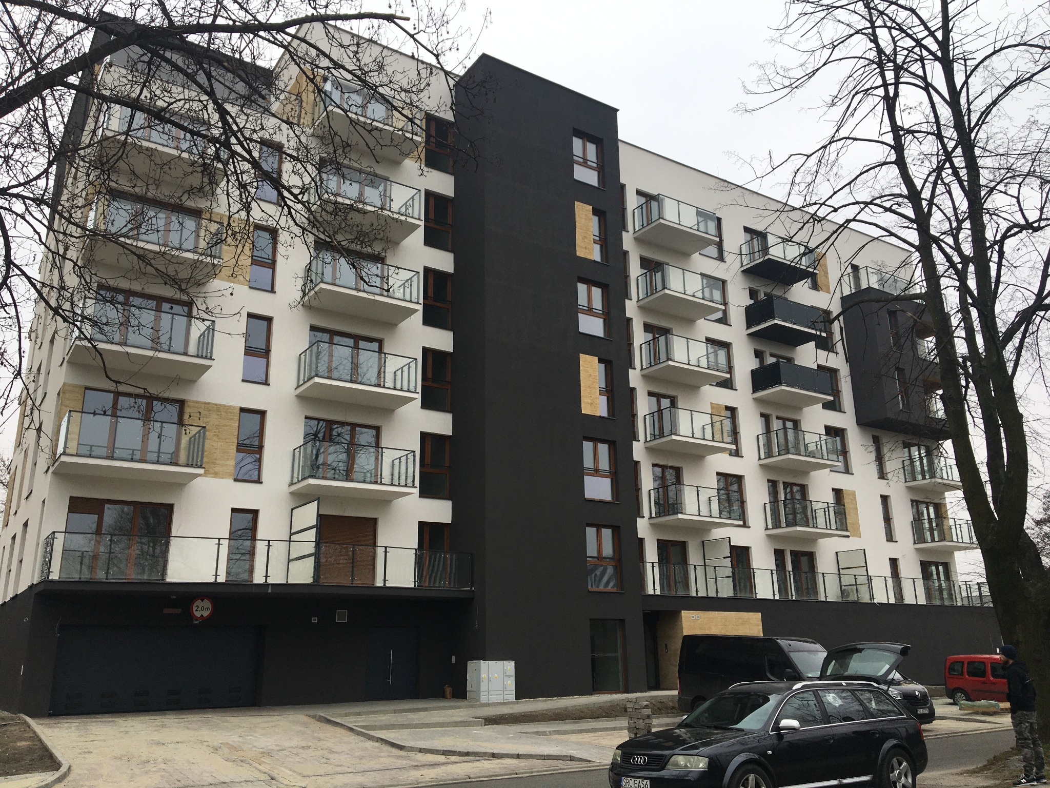 Nowoczesny budynek mieszkalny z balkonami zabezpieczonymi szklanymi balustradami, widok z poziomu ulicy, samochody zaparkowane przed budynkiem.