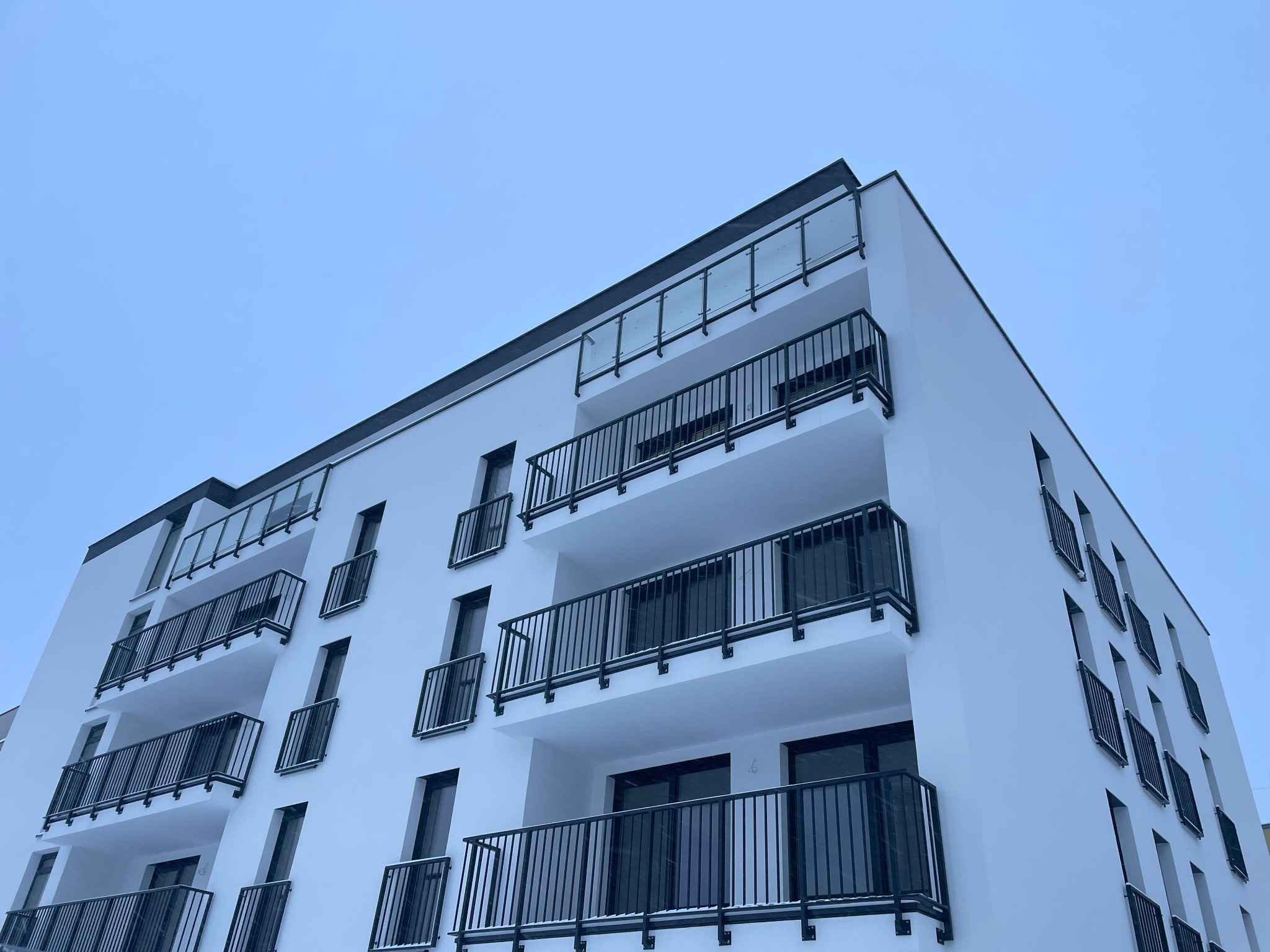 Nowoczesny budynek mieszkalny z białymi elewacjami i balkonami z czarnymi, metalowymi balustradami, sfotografowany z dołu pod kątem, na tle jasnego nieba.