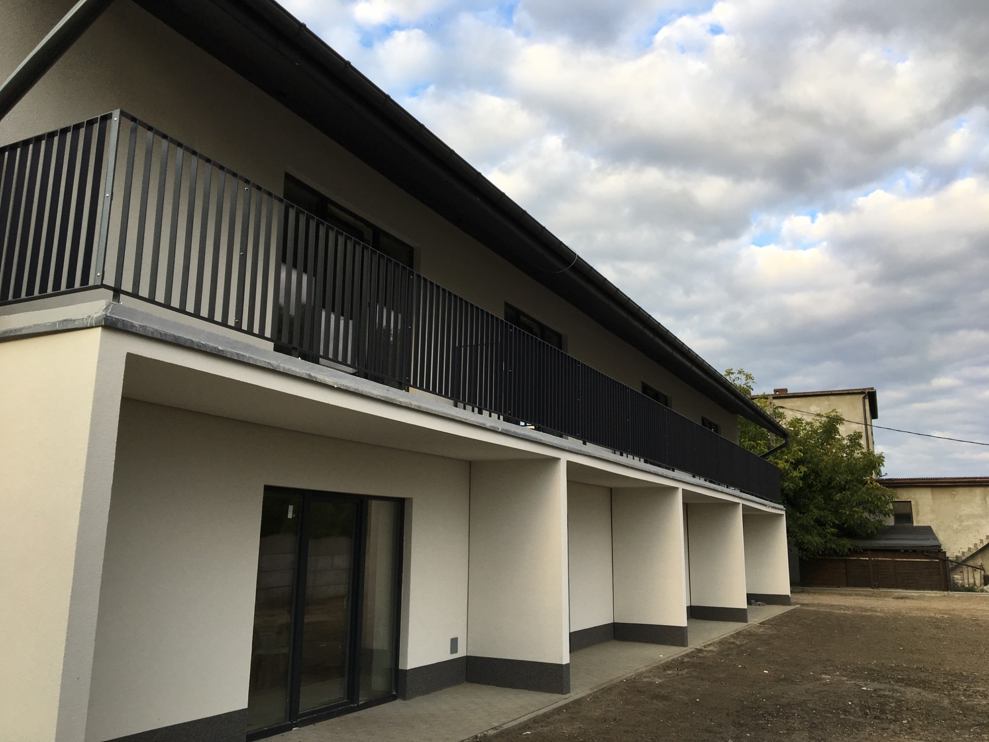 Nowoczesny budynek z długim balkonem zabezpieczonym czarną, pionową balustradą metalową; widok z dołu na tle pochmurnego nieba.
