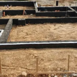 Fundamenty domu jednorodzinnego z hydroizolacją i wystającymi prętami zbrojeniowymi, otoczone piaskiem i drewnianymi stemplami szalunkowymi.