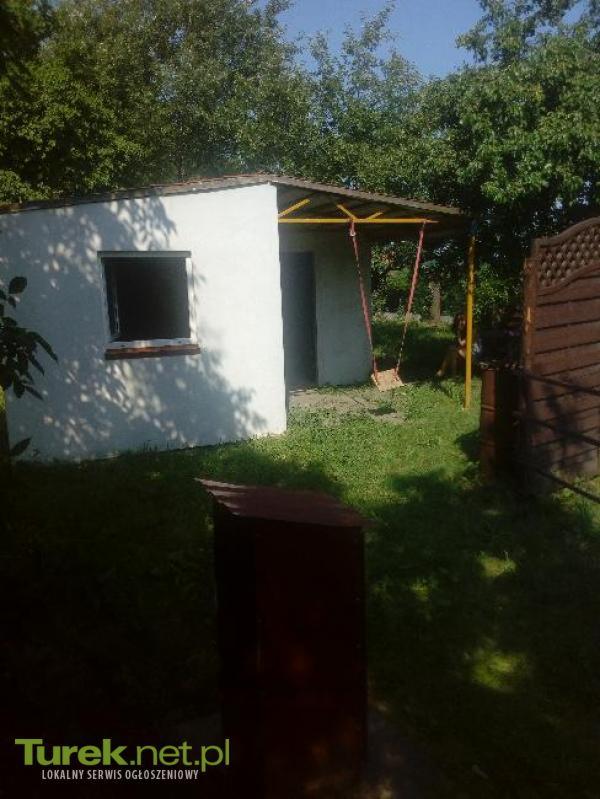 Mały, biały budynek z płaskim dachem i zadaszeniem na słupach, prawdopodobnie domek narzędziowy lub letniskowy, z huśtawką na tle zieleni, w słoneczny dzień.