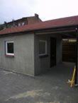 Niewielki, parterowy budynek z szarą elewacją, białym obramowaniem okna i czerwonym dachem, przylegający do niego zadaszony garaż.