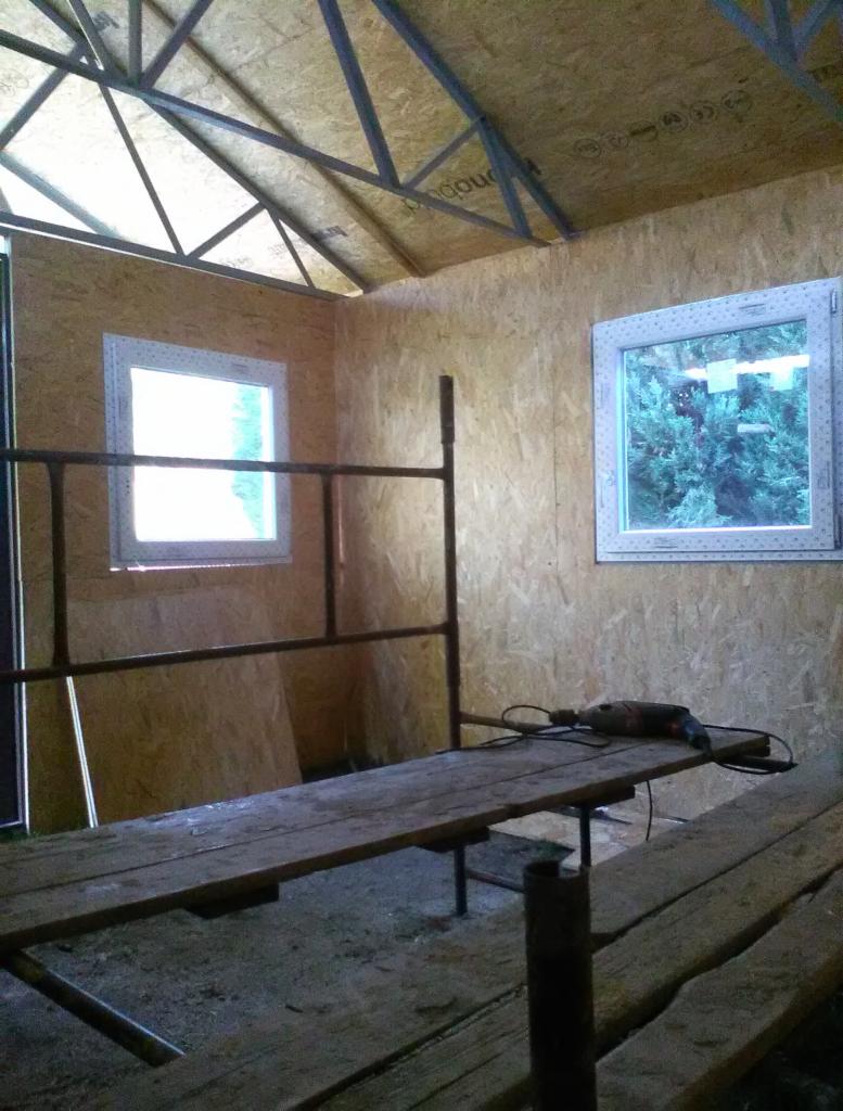 Wnętrze w trakcie budowy z płyt OSB, widoczne okna z białymi ramami, metalowa konstrukcja dachu i wiertarka na drewnianym rusztowaniu.