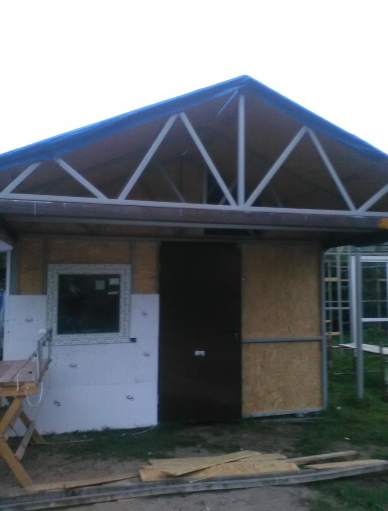 Budowa małego budynku z dachem dwuspadowym pokrytym niebieską plandeką, widoczne płyty OSB i okno z białymi ramami, na zewnątrz trawa i resztki materiałów budowlanych.