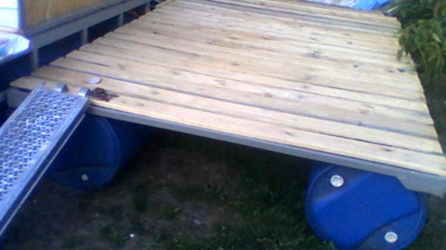 Drewniana platforma na plastikowych beczkach z metalową rampą, widok z boku, w tle zieleń.