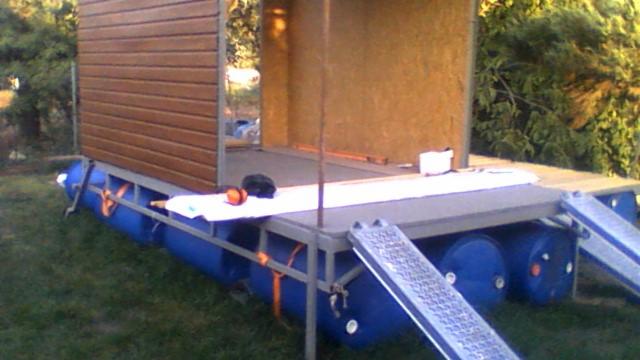 Niedokończona konstrukcja pływająca z elementami drewnianymi, oparta na niebieskich beczkach, z widocznymi rampami wejściowymi i akcesoriami budowlanymi na pokładzie.