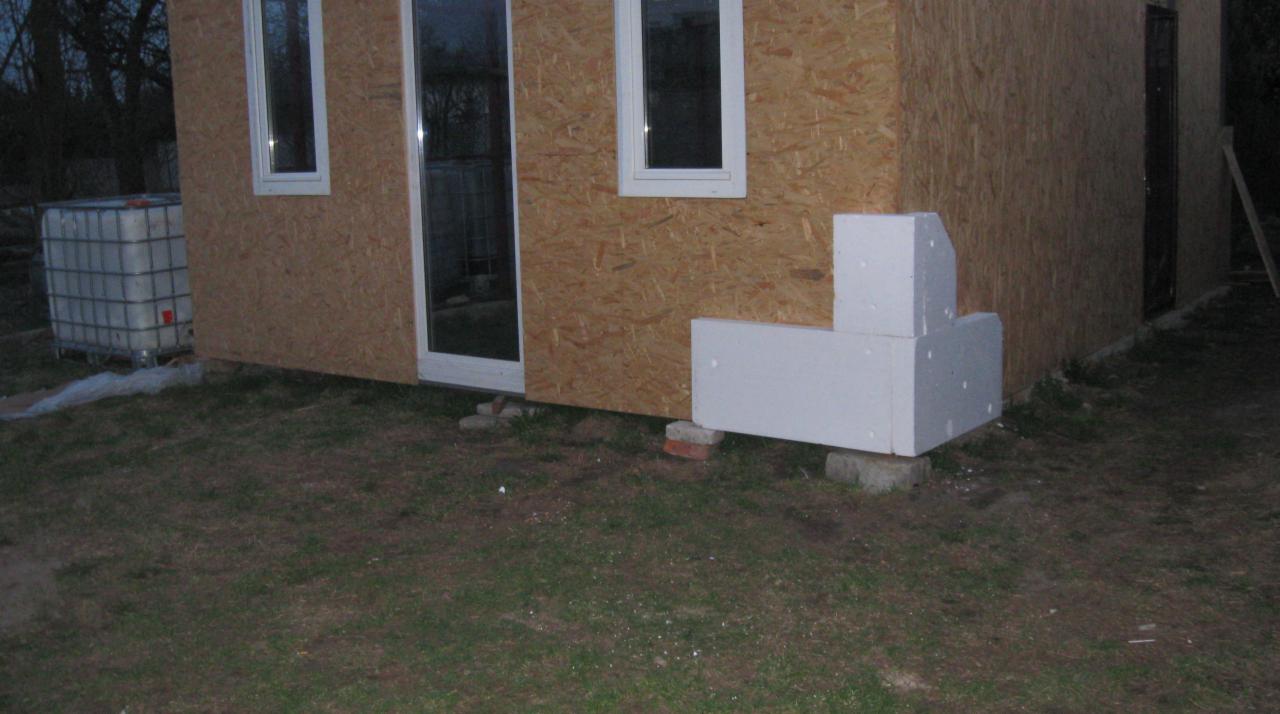 Surowy stan budynku szkieletowego z płyt OSB, widoczne okna, drzwi, białe płyty styropianowe przy fundamentach i zbiornik na wodę w metalowej klatce z tyłu.