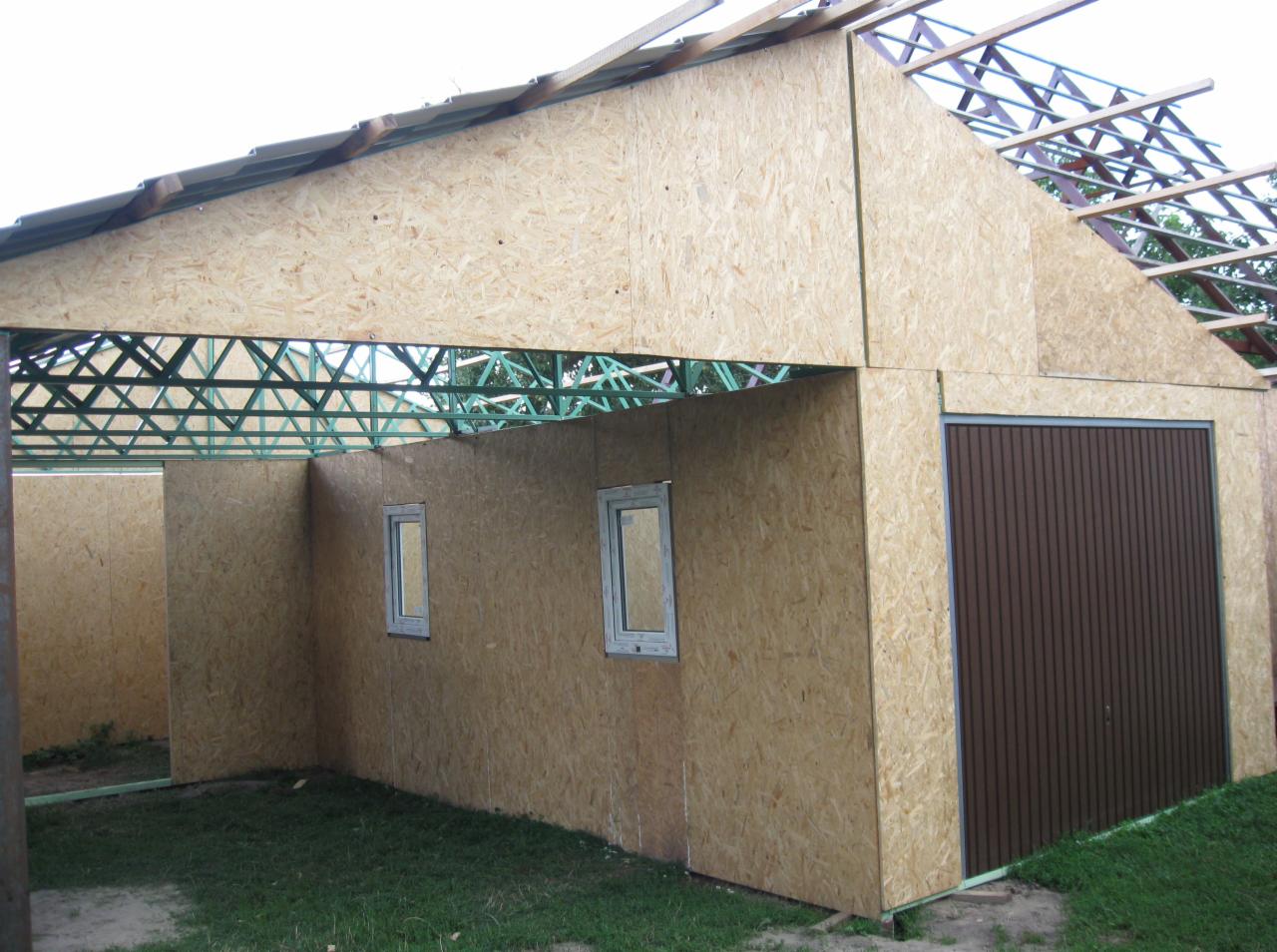 Surowa konstrukcja garażu z płyty OSB z brązową bramą segmentową, widoczne elementy konstrukcji dachu i zielona stalowa rama.