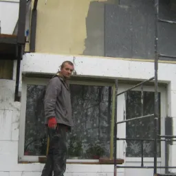 Ocieplanie elewacji budynku styropianem, widoczny pracownik na rusztowaniu w czerwonych rękawicach, okno z firanką w tle, częściowo otynkowana ściana.