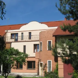 Dwupiętrowy dom z beżową elewacją, czerwonym dachem i okrągłą wieżyczką z cegły, otoczony zielenią i drzewami w słoneczny dzień.