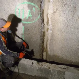 Pracownik w pomarańczowej kamizelce uszczelnia pęknięcie w betonowej ścianie, z której wycieka woda. Na ścianie zielony okrąg z napisem.