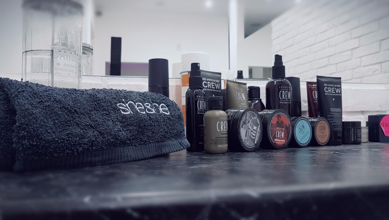 Różnorodność produktów marki American Crew na czarnym blacie, obok złożony, ciemny ręcznik z wyhaftowanym napisem, w tle biała ceglana ściana.