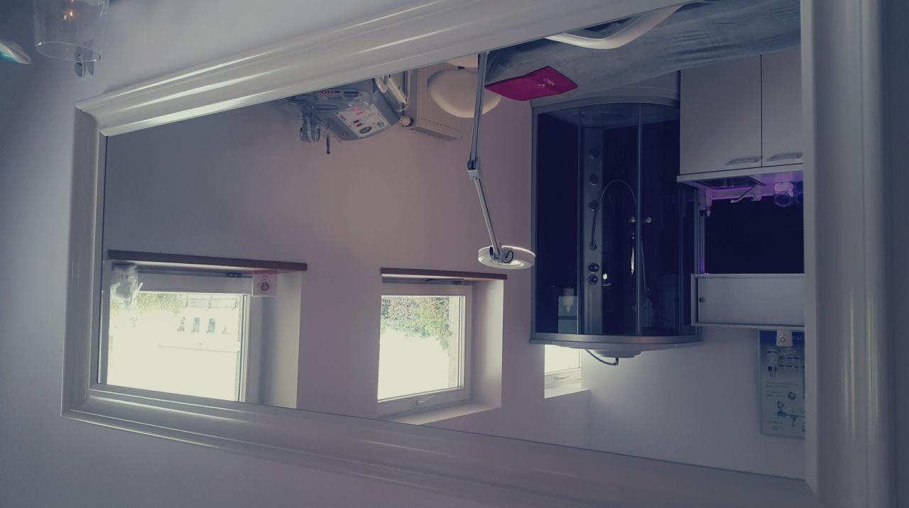 Odbicie w lustrze: Nowoczesna kabina prysznicowa z hydromasażem, białe szafki łazienkowe z fioletowym podświetleniem i okna z widokiem na zewnątrz.