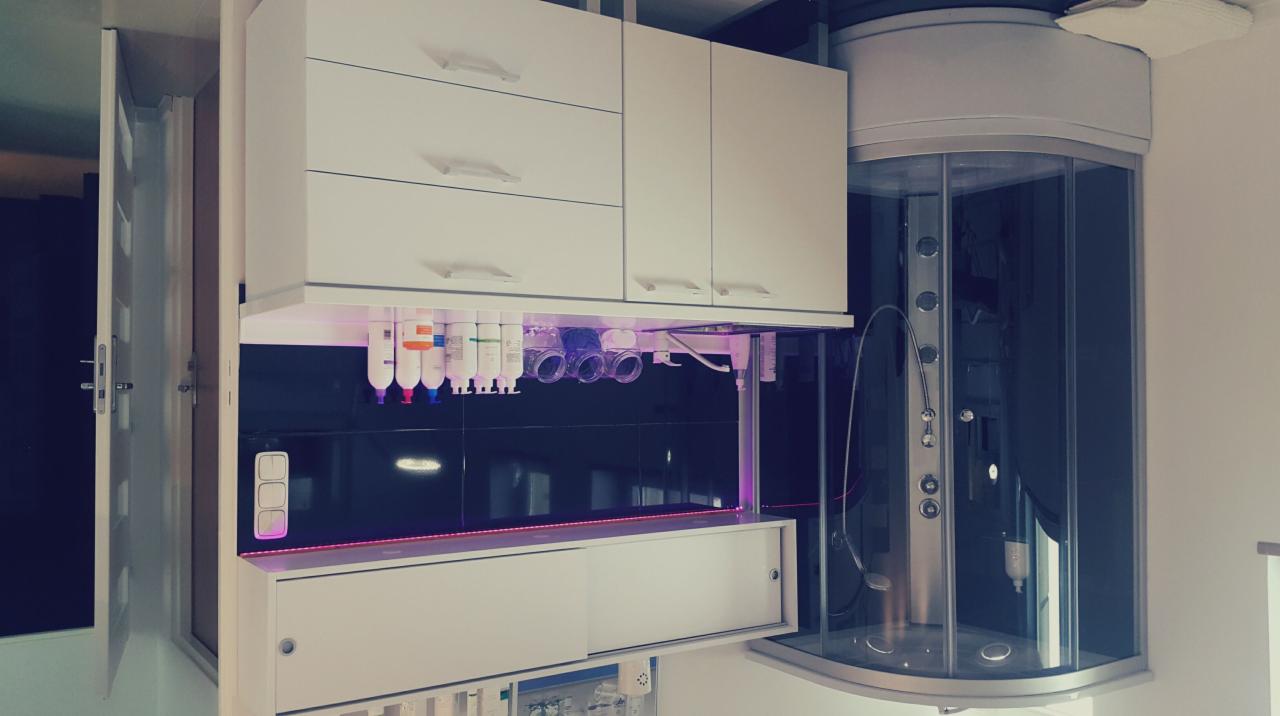 Wyposażenie salonu kosmetycznego: białe szafki wiszące z kosmetykami podświetlonymi fioletowym światłem, czarne płytki na ścianie i kabina prysznicowa z hydromasażem w tle.