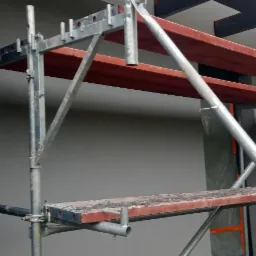 Szare ściany budynku w trakcie prac wykończeniowych, widoczne rusztowanie z drewnianymi pomostami, na ścianie widoczne fragmenty elewacji w kolorze czarnym.