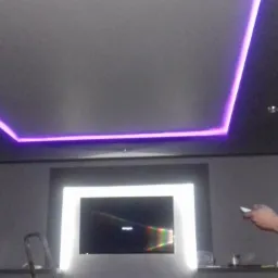 Szare wnętrze pokoju z podświetlanym fioletowym oświetleniem LED na suficie, podświetlonym telewizorem i fragmentem drabiny.