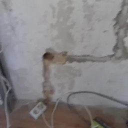 Przygotowanie ściany pod gładzie: wycięte bruzdy pod instalacje elektryczne i rury, widoczne gniazdko elektryczne, przewody i narzędzia na drewnianej podłodze.