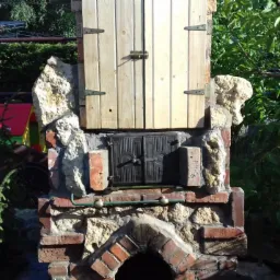 Ogród z murowanym grillem z kamienia i cegły, z drewnianymi i metalowymi drzwiczkami, otoczony zielenią.