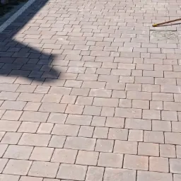 Ułożona nawierzchnia z prostokątnej kostki brukowej w odcieniach brązu, widoczny fragment korytka odwadniającego z białym obramowaniem i studzienka kanalizacyjna z żółtą rączką narzędzia obok.