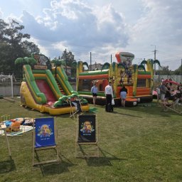 Wyspa Uśmiechu Jadwiga Surma - Dwa dmuchańce w stylu safari (zjeżdżalnia i zamek) na trawie, otoczone przez ludzi. Na pierwszym planie dwa leżaki z logo 'Wyspa'. Słoneczny dzień.