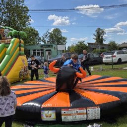Wyspa Uśmiechu Jadwiga Surma - Dzieci bawią się na dmuchanym rodeo byku i zjeżdżalni na świeżym powietrzu podczas imprezy plenerowej w słoneczny dzień. W tle budynek i zaparkowane samochody.