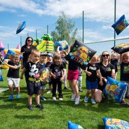 Wyspa Uśmiechu Jadwiga Surma - Grupa dzieci na trawie trzymająca poduszki z logo 'Wyspa', w tle dmuchany zamek i animatorzy, słoneczny dzień.