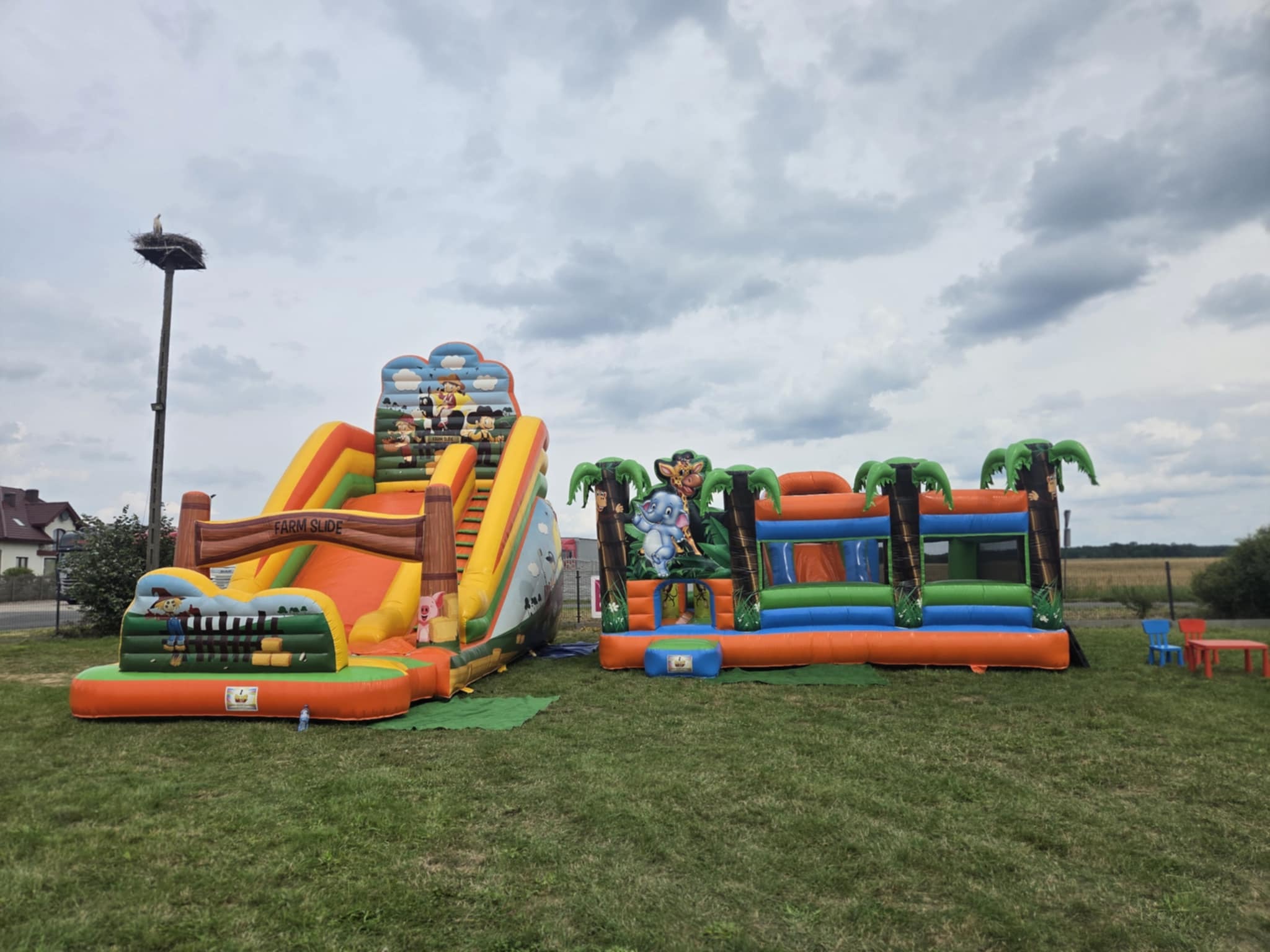 Dwa dmuchane zamki na trawie: jeden ze zjeżdżalnią z motywem farmy, drugi z motywem dżungli. W tle widoczne budynki i pochmurne niebo. Na słupie gniazdo bocianie.