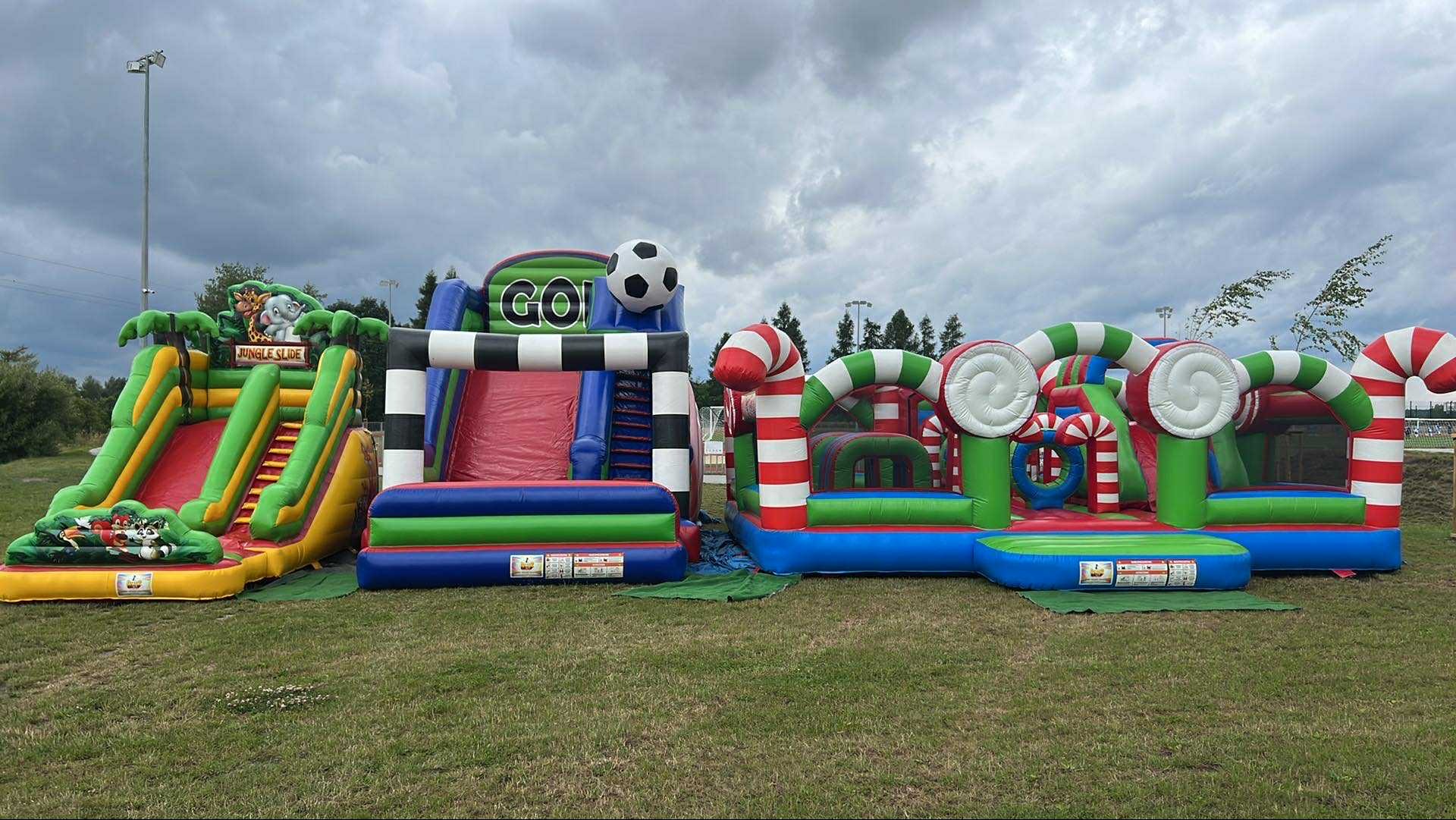 Kompleks dmuchańców: zjeżdżalnia dżungla, tor przeszkód z piłką i świąteczny plac zabaw z lizakami. Kolorowe atrakcje na trawie pod pochmurnym niebem.