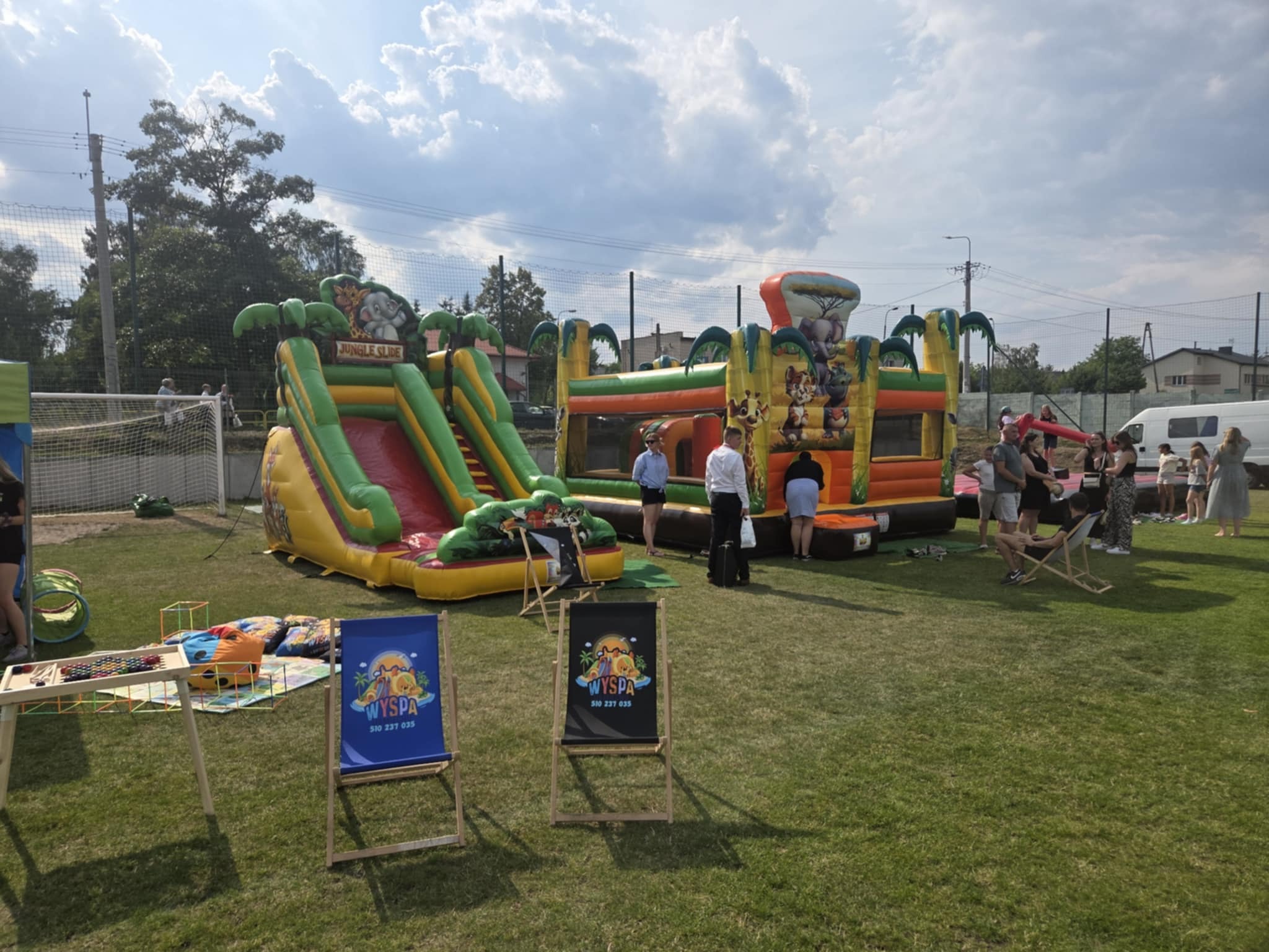 Dwa dmuchańce w stylu safari (zjeżdżalnia i zamek) na trawie, otoczone przez ludzi. Na pierwszym planie dwa leżaki z logo 'Wyspa'. Słoneczny dzień.