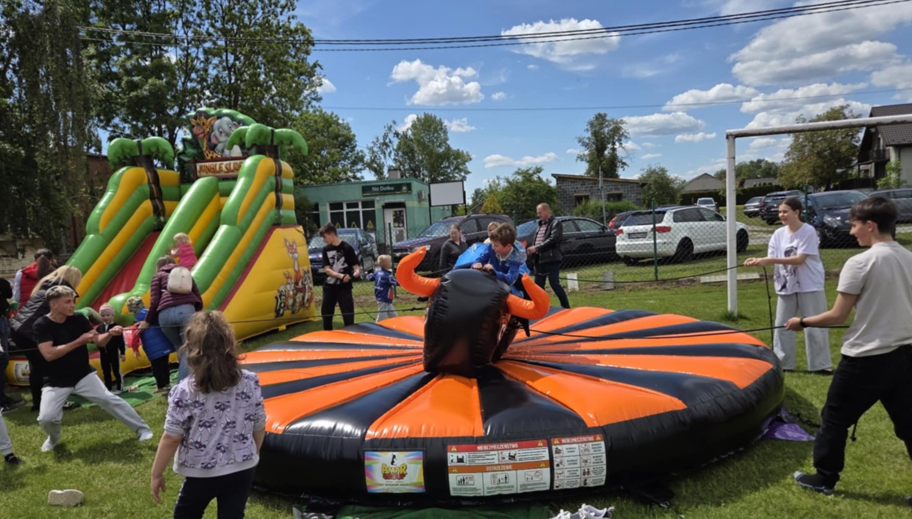 Dzieci bawią się na dmuchanym rodeo byku i zjeżdżalni na świeżym powietrzu podczas imprezy plenerowej w słoneczny dzień. W tle budynek i zaparkowane samochody.