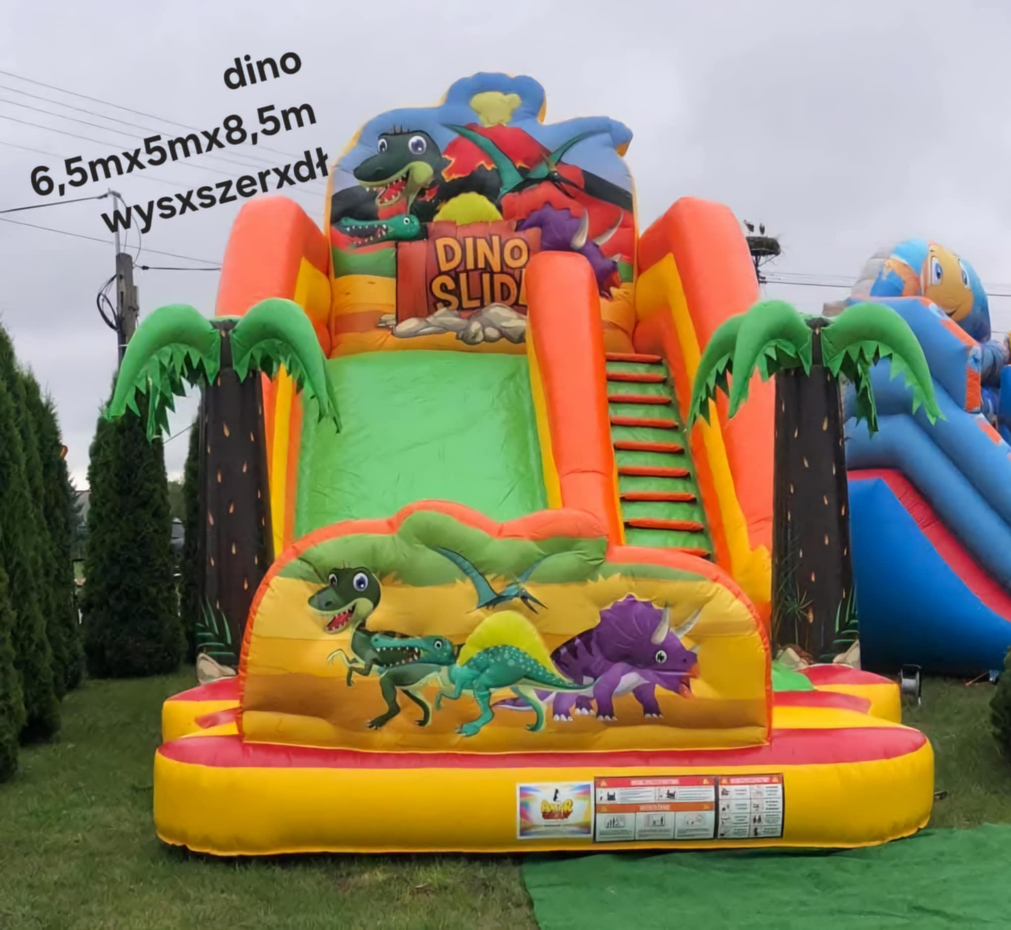 Kolorowy dmuchany zamek ze zjeżdżalnią Dino Slide, z motywem dinozaurów i palm, ustawiony na trawie. Widoczny inny dmuchaniec w tle.