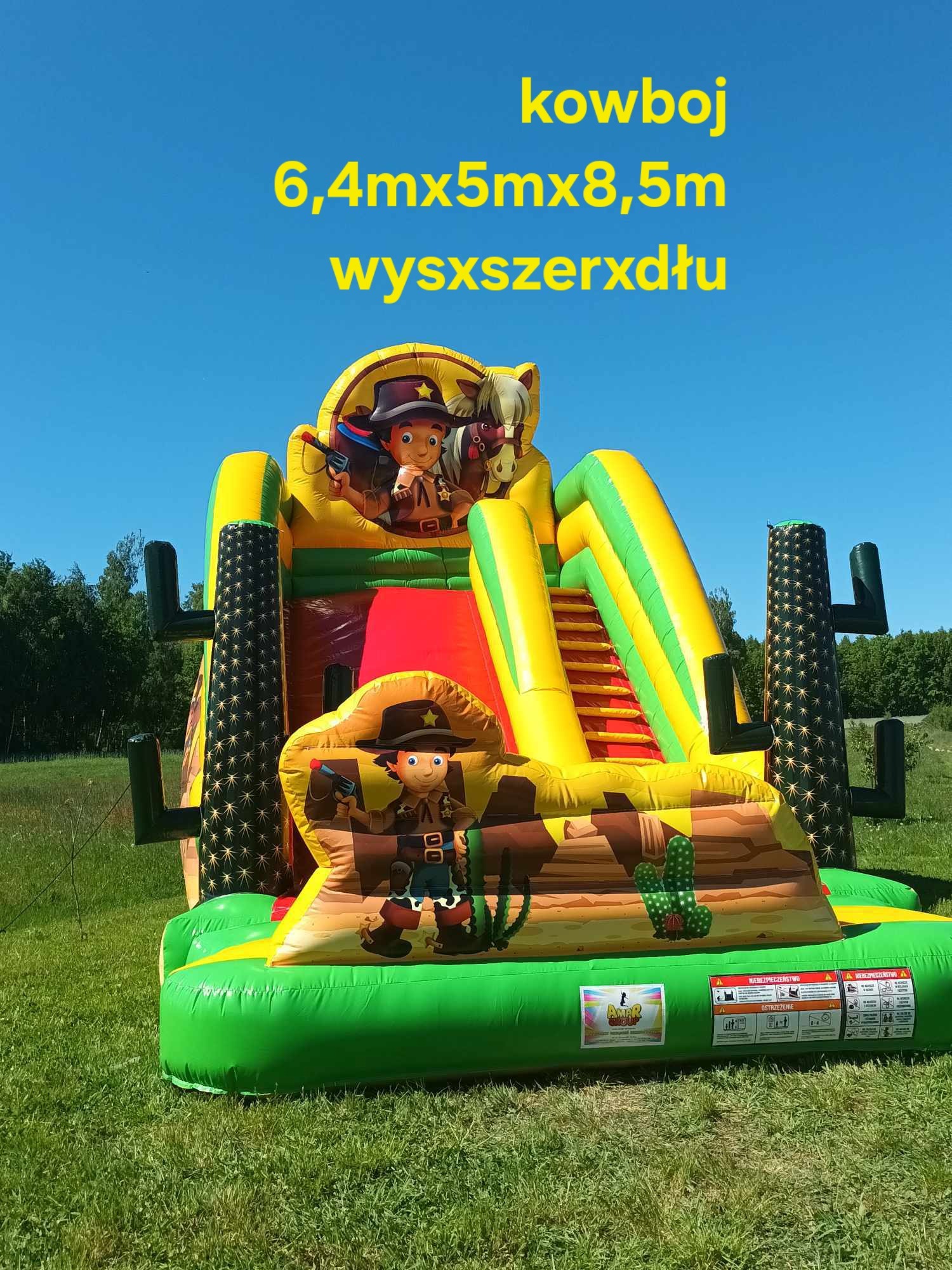 Kolorowy dmuchany zamek w stylu kowbojskim na zielonej trawie, z motywami kaktusów i postaci kowbojów. Widoczne schody i zjeżdżalnia. W tle drzewa i błękitne niebo.