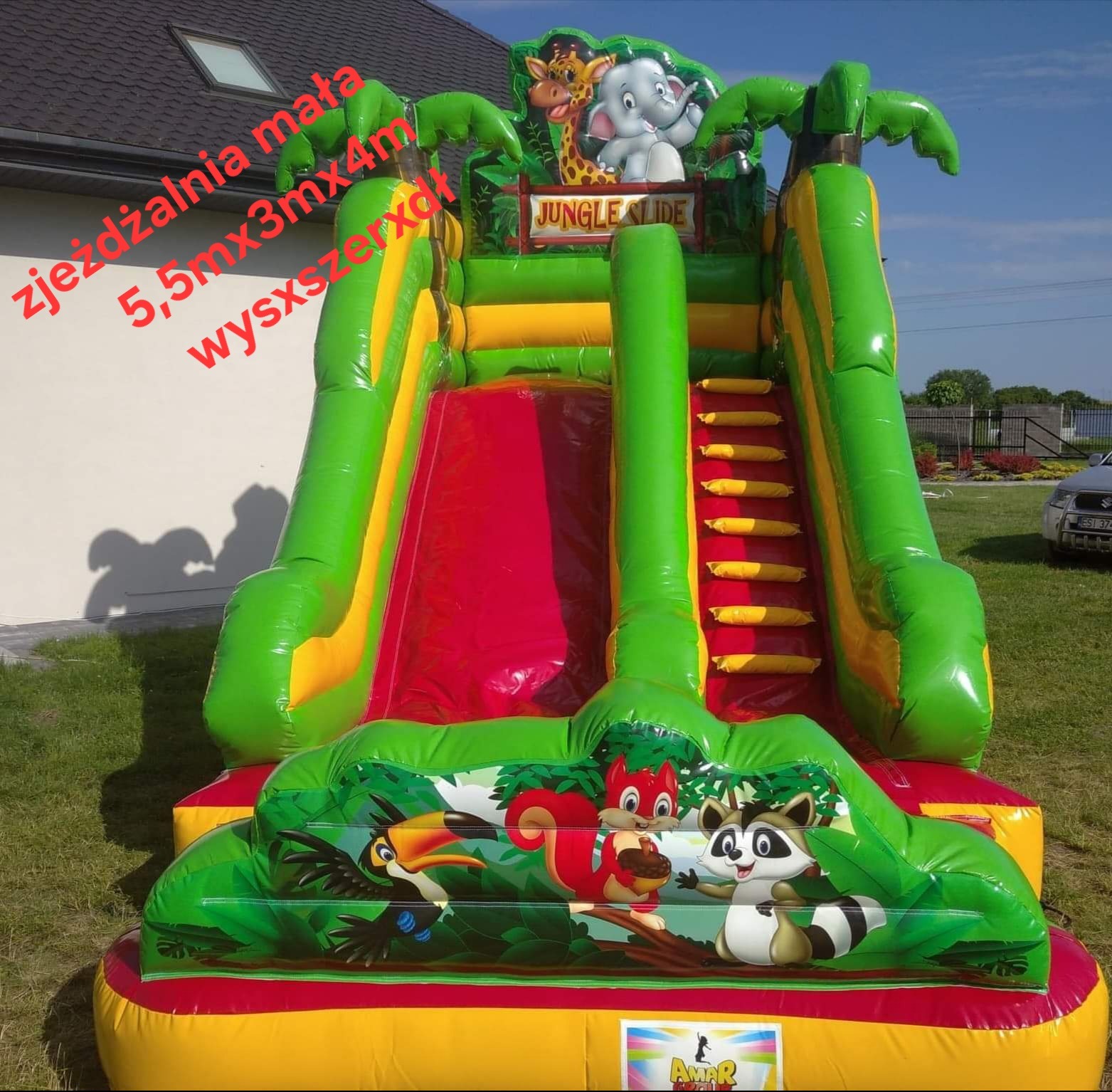 Kolorowa, dmuchana zjeżdżalnia z motywem dżungli, z żyrafą, słoniem i palmami. Widoczne schody i ślizg. Zjeżdżalnia stoi na trawie, w tle dom i niebieskie niebo.
