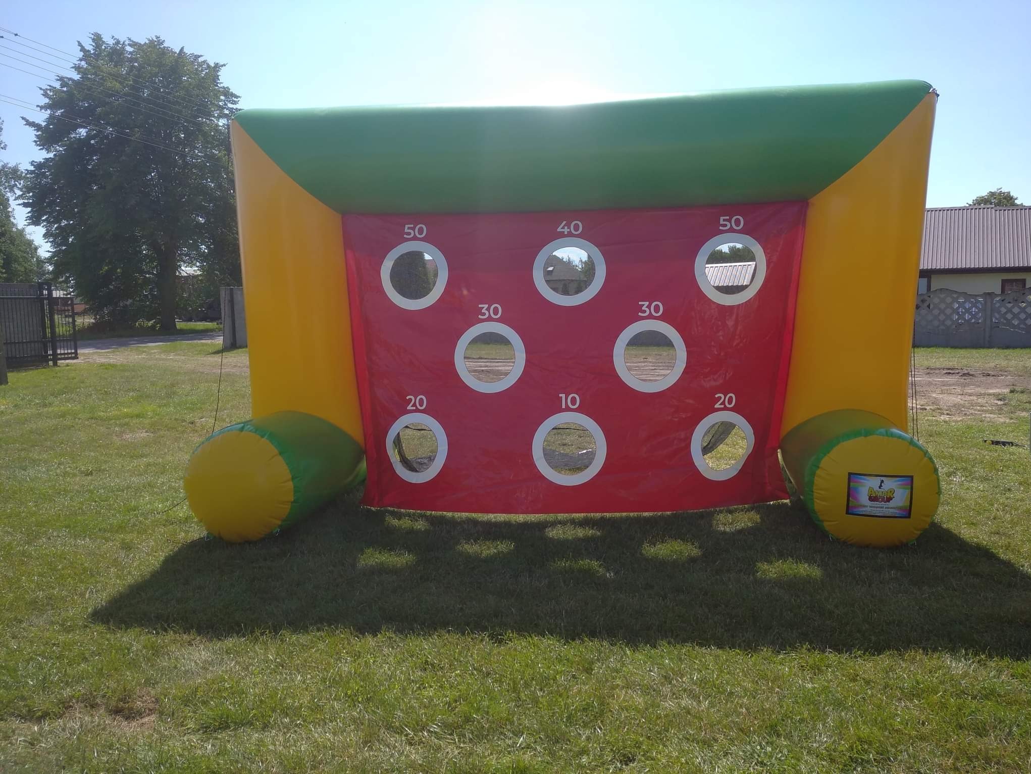 Kolorowa, dmuchana bramka celnościowa z otworami punktowanymi (10-50) na trawie, idealna na imprezy plenerowe i eventy dla dzieci. Żółto-zielona konstrukcja z czerwonym panelem.