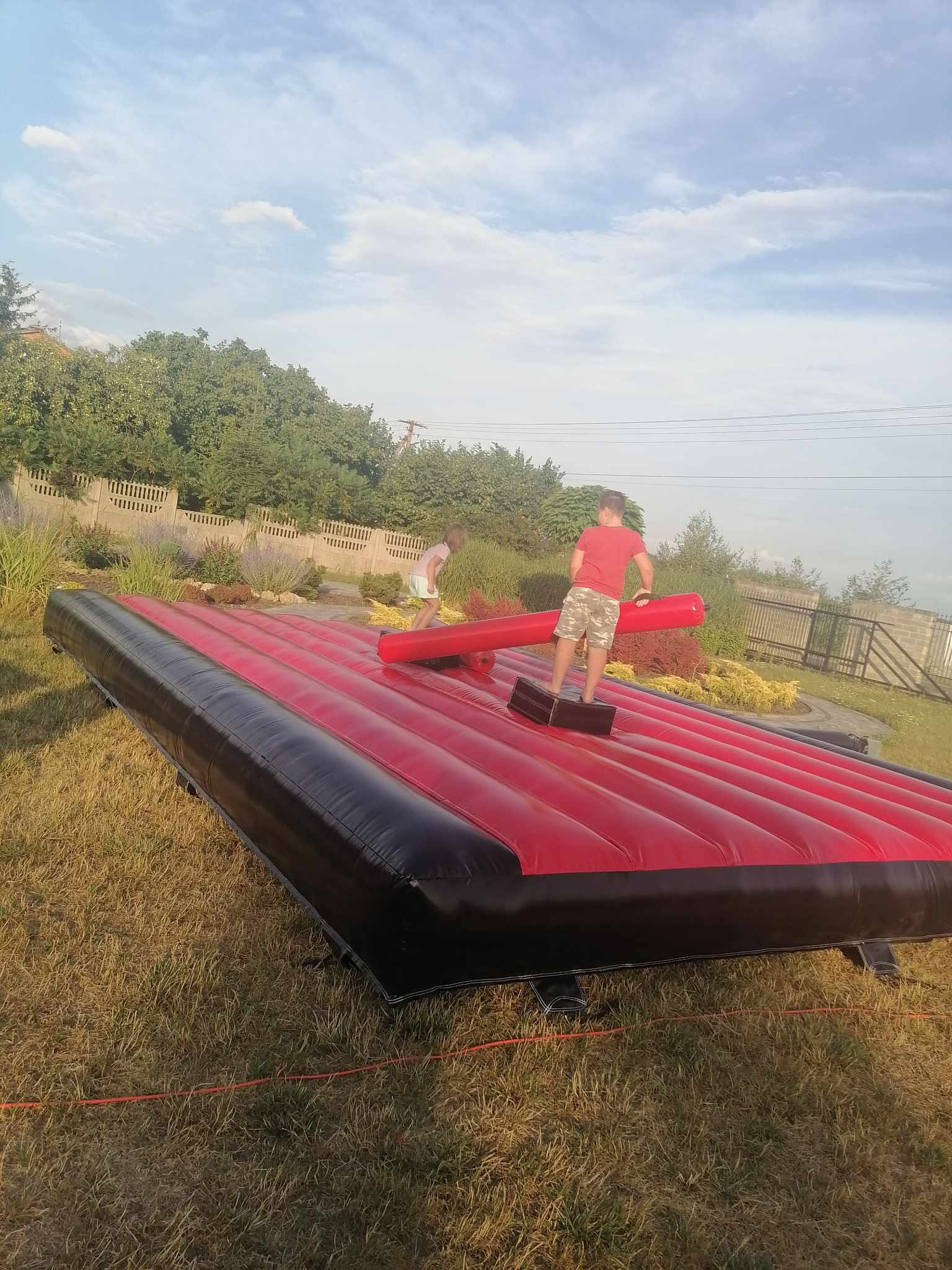 Dzieci na dmuchanym torze przeszkód z elementami do walki, ustawionym na trawie w ogrodzie. W tle drzewa i niebo.