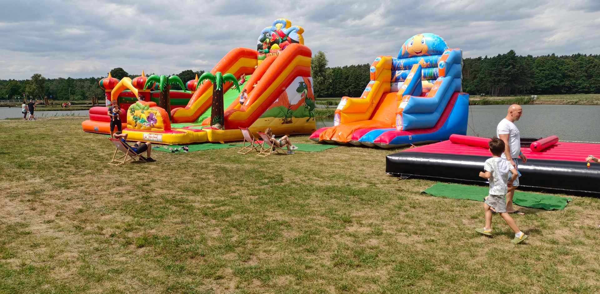 Kolorowe dmuchańce z motywami dinozaurów i ryb na trawie, obok jeziora. W tle drzewa i pochmurne niebo. Widoczni ludzie odpoczywający na leżakach i dziecko.
